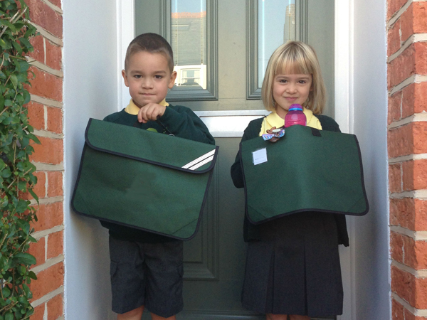 A boy and a girl stand in front of a house door holding up their matching school bags