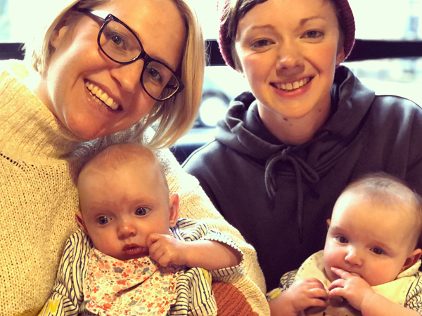 Two women sit smiling at the camera holding twin babies