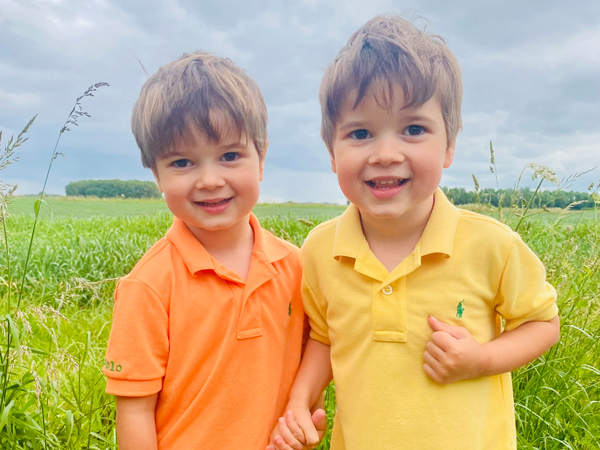 A set of twins around five years old stand in front of a field of wheat smiling at the camera