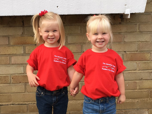 Two girls in matching red t-shirts stand in front of a wall smiling at the camera