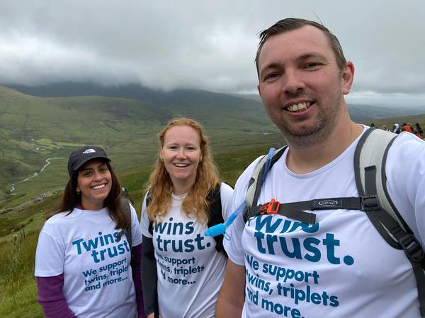 Three adults wearing Twins Trust t-shirts pose for a selfie with a mountainous background