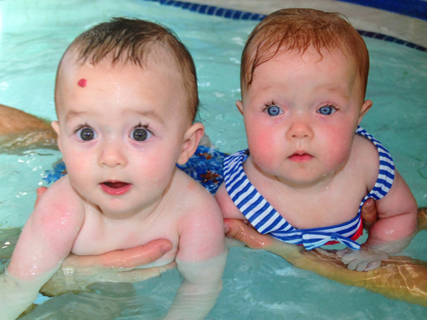 Picture showing two twins being held in a swimming pool, the twins look similar but not identical and one has blue eyes whilst the other has brown eyes