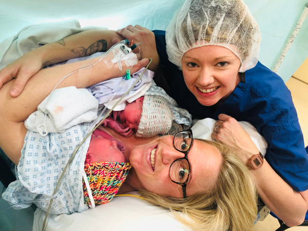 A woman lies on a bed in a hospital grown holding a baby whilst another woman sits next to her