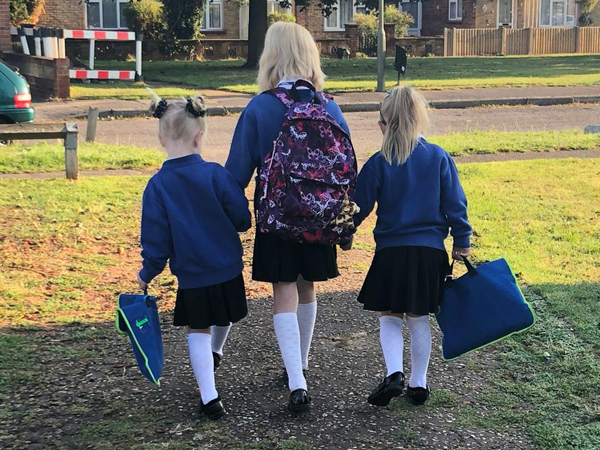Three children dressed in identical school uniforms walk away from the camera whilst holding hands with a taller child in the middle of two shorter children
