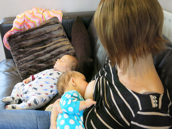 The camera looks down on a mother who is breastfeeding one baby whilst another sits on the sofa next to her