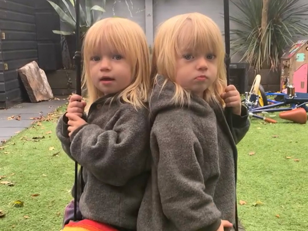 Twin girls stand back-to-back in a garden with their faces turned towards the camera